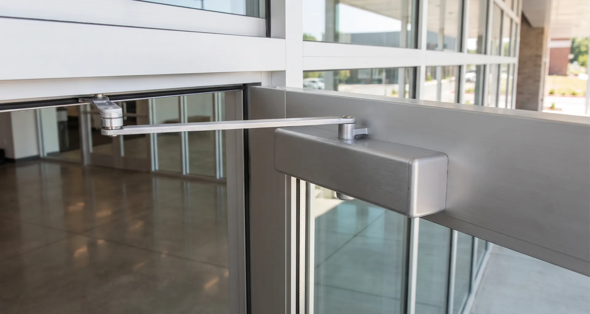 A close-up of a silver commercial door closer unit attached to the top right corner